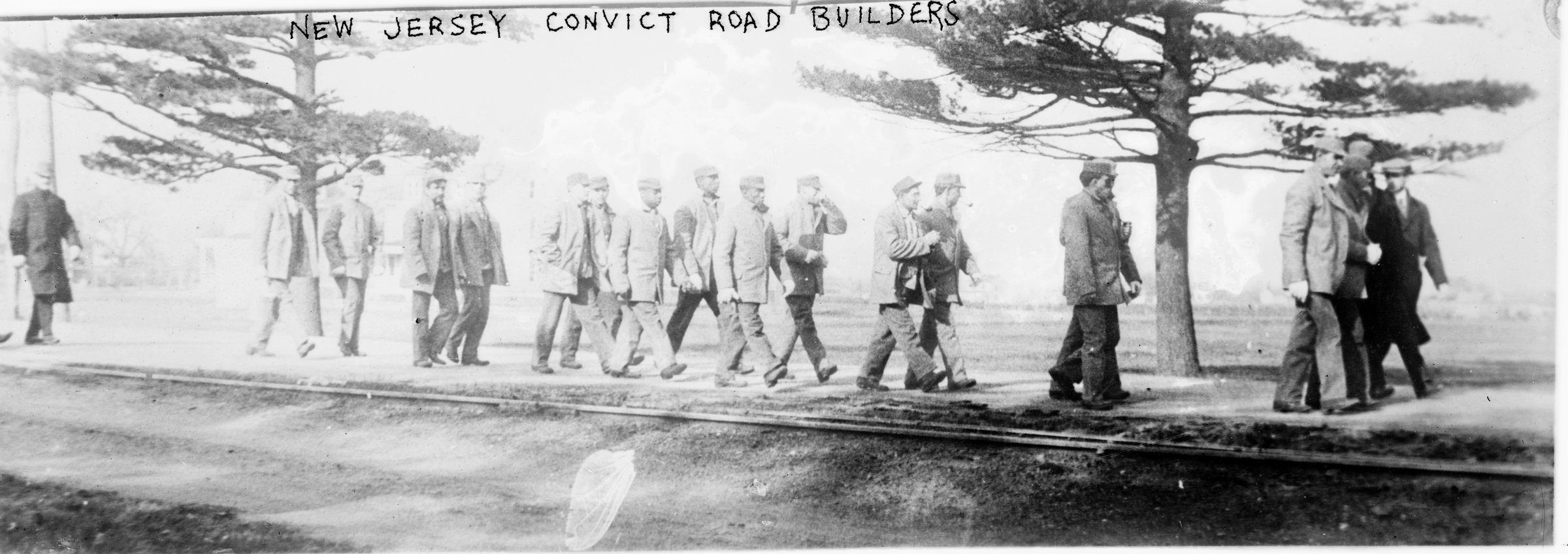 A line of convicts in work clothes marching along a road under bare trees, captioned on the negative New Jersey Convict Road Builders